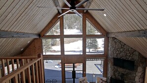 A spacious room with a high ceiling, large windows offering a view of a snowy landscape, a stone fireplace, and a wooden staircase. - "Peace Like a River" mountain cabin on river - Red River, NM. Hot tub optional. (Red River)
