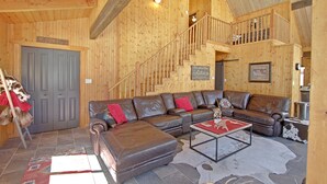 A spacious living room with a large brown leather sectional sofa, a coffee table, and a staircase leading to an upper level. - "Peace Like a River" mountain cabin on river - Red River, NM. Hot tub optional. (Red River)
