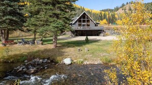 A lodge with a stone foundation, a wooden deck, and a view of a mountain range. - "Peace Like a River" mountain cabin on river - Red River, NM. Hot tub optional. (Red River)