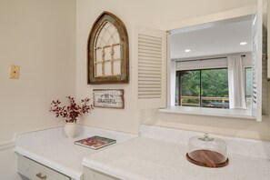 A kitchen with a white countertop, a decorative window with a view of greenery, a white vase with red leaves, and a wooden serving tray. - K K’s Hideaway—pond/pool table 

 (Hattiesburg)