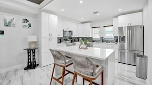 Una cocina moderna con armarios blancos, una isla central con dos sillas de mimbre, electrodomésticos de acero inoxidable y suelo de mármol.