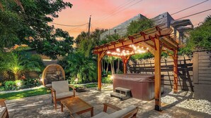 Un área de patio al aire libre con mobiliario de madera, un jacuzzi y una pérgola con luces colgantes.