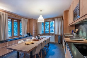 A modern kitchen with wooden cabinets, a dining table with chairs, a hanging light fixture, and a view of trees through large windows.