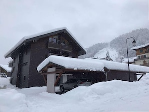A two-story chalet with a covered garage and a car parked inside. Snow covers the roof and ground.