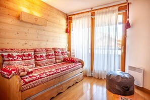 A cozy room with a wooden log wall, a sofa with patterned cushions, a window with sheer curtains, and a radiator.