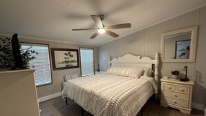 A bedroom with a bed, bedside table, nightstand, ceiling fan, and a large window with blinds.