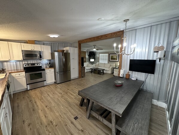 A modern kitchen with stainless steel appliances, a wooden dining table, and a living area with a sofa and TV.