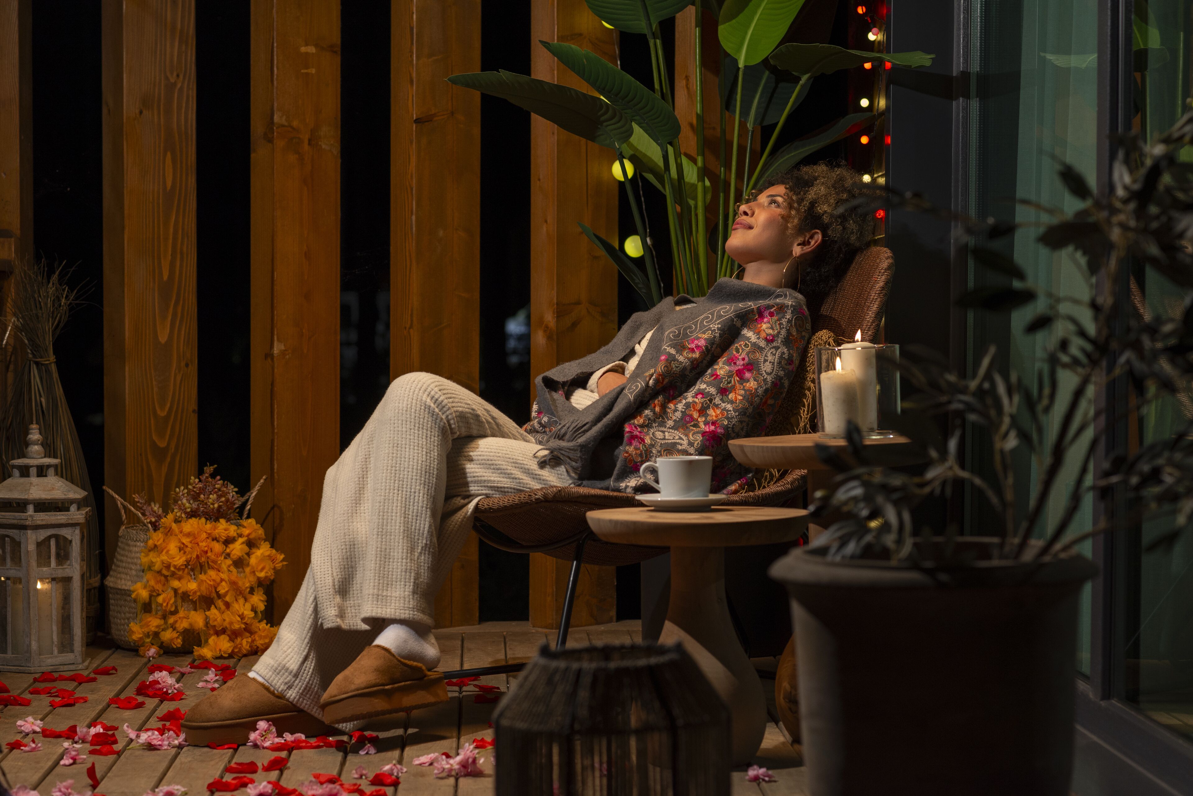 A person reclining in a chair with a cup and candle, surrounded by plants and flowers.