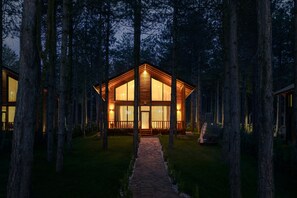 Een houten hut met grote ramen, omringd door hoge bomen en een pad van stenen dat naar de ingang leidt.