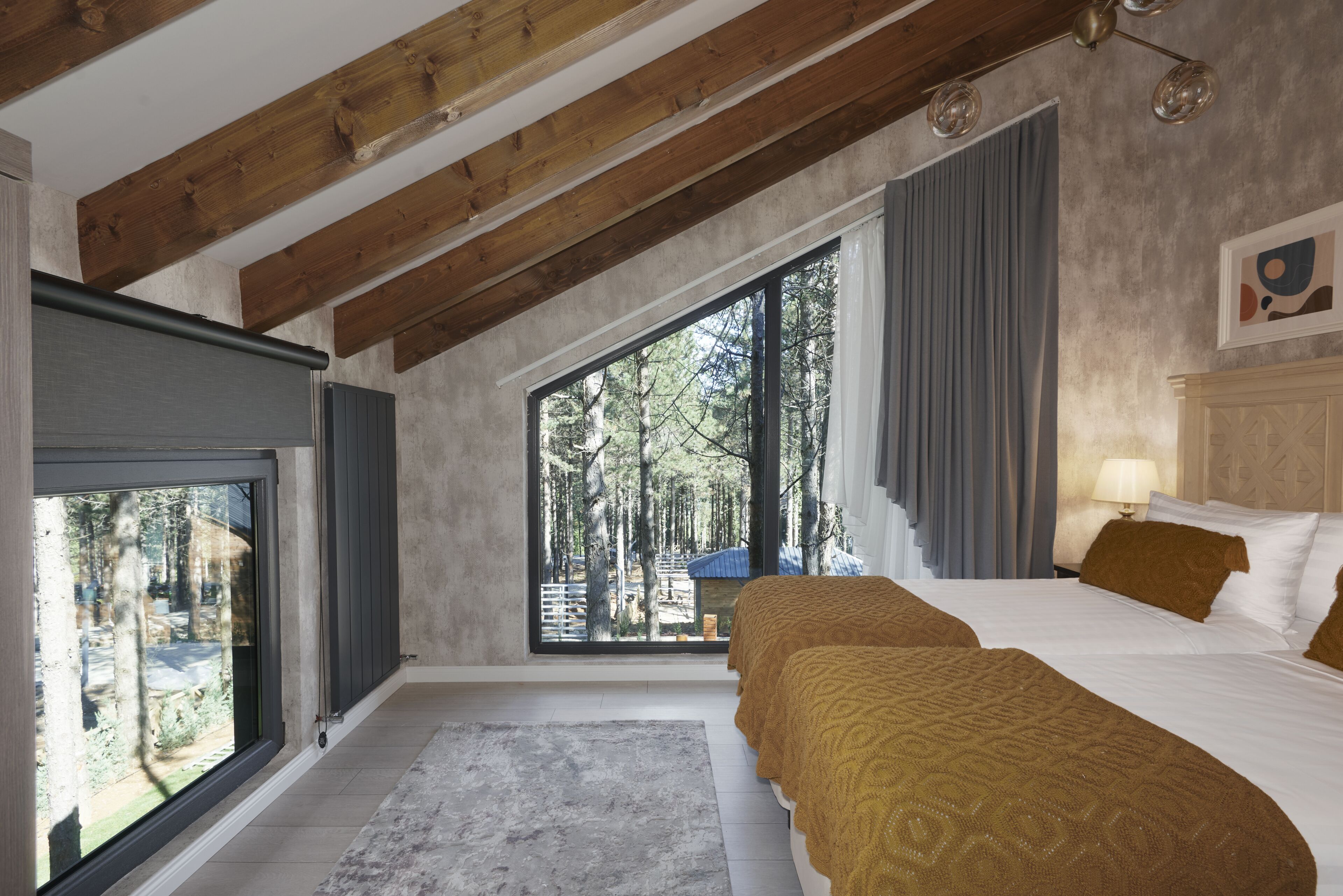 A bedroom with a large window, wooden beams, a bed with a patterned blanket, and a view of trees.
