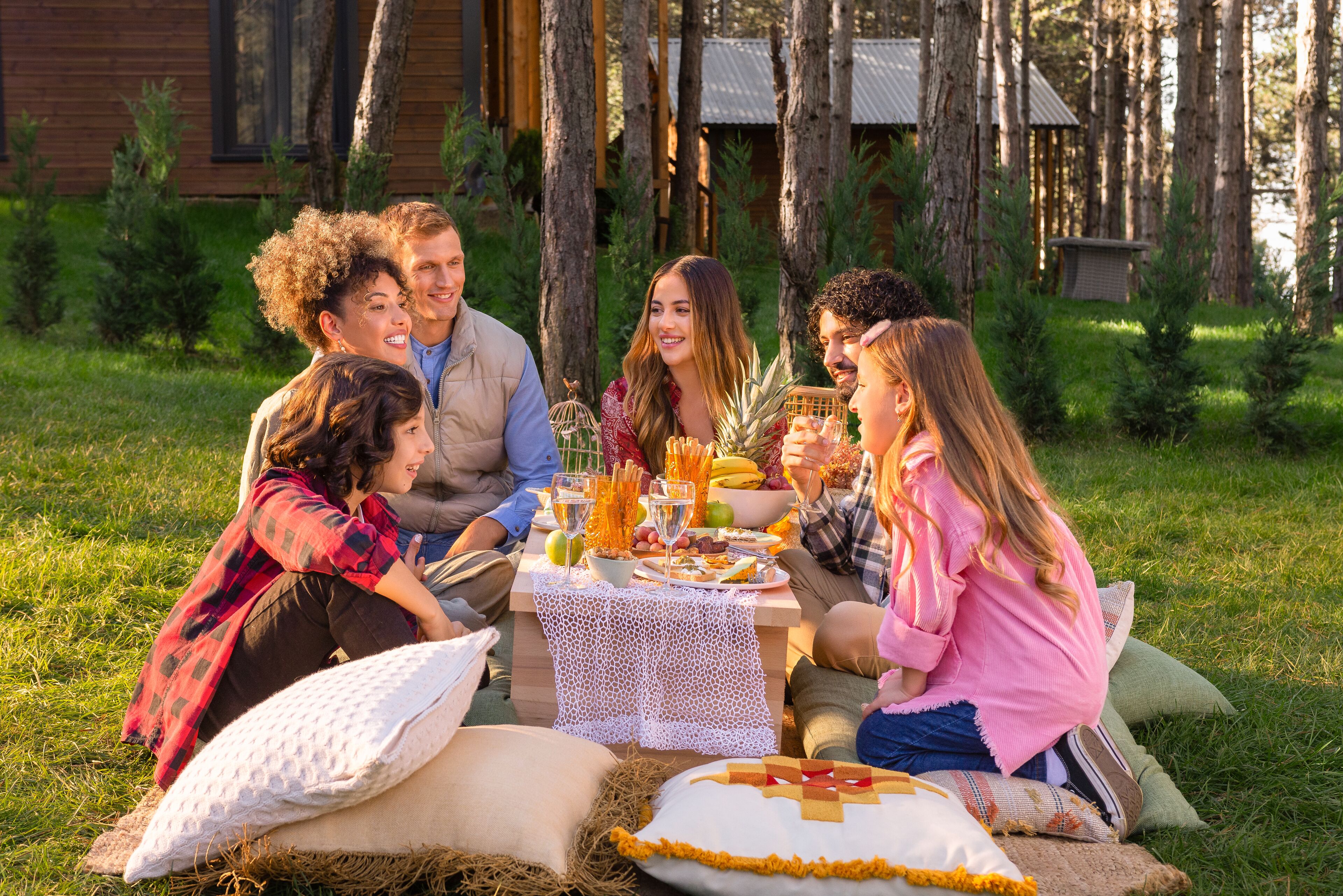 A group of people sitting at an outdoor table with a picnic setup, surrounded by trees and a cabin in the background.
