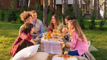 A group of people sitting at an outdoor table with a picnic setup, surrounded by trees and a cabin in the background.