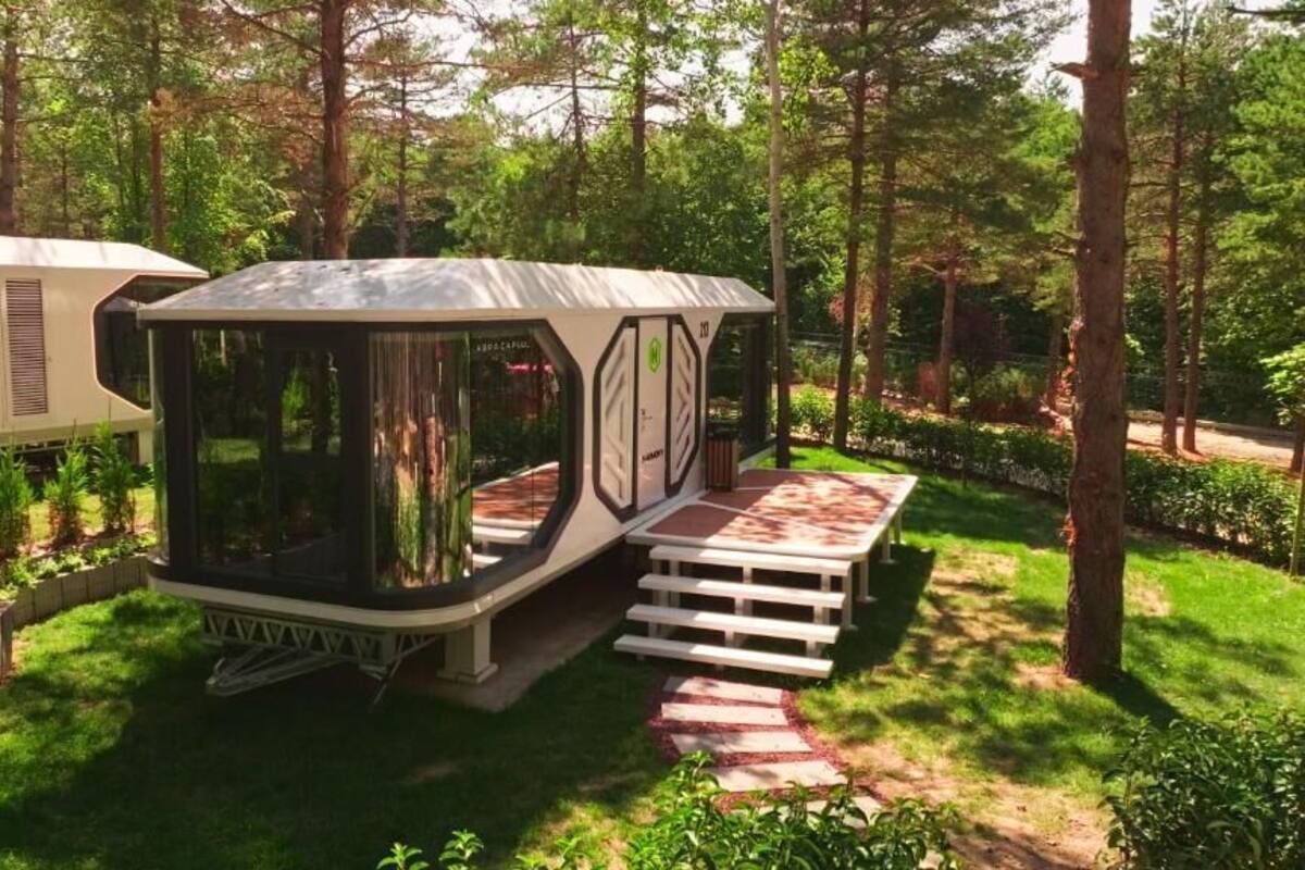 A modern, compact RV with a glass enclosure, situated in a lush, green forest setting.