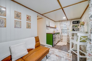 Un salón acogedor con un sofá marrón y una almohada blanca, una pequeña cocina con un microondas y un armario verde, y un comedor con una silla blanca.