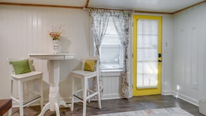 A room with a yellow door, white walls, a small table with two chairs, a vase with flowers, and a window with floral curtains. - Wilmington Area Studio ~ 4 Mi to Downtown! (Leland)