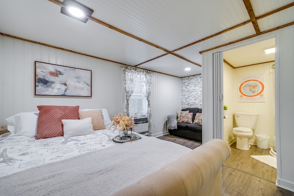 Un dormitorio con cama, mesita de noche con lámpara, ventana con cortinas, una obra de arte colgada en la pared, y un baño visible a través de una puerta abierta.