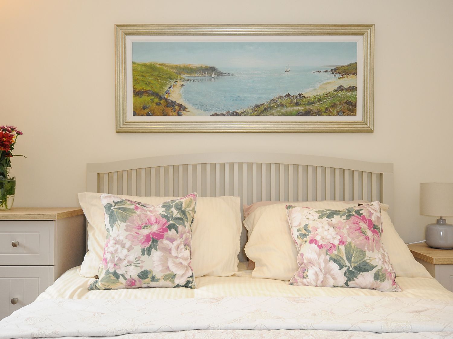 Un dormitorio con una cama, almohadillas florales, una mesita de noche con lámpara, un cuadro enmarcados con una escena costera y un florero con flores.