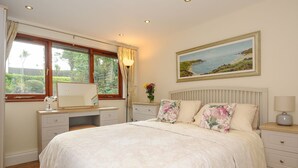 A bedroom with a large bed, a desk with a mirror, a nightstand, and a painting of a coastal landscape. - Little Trevallan (Looe)