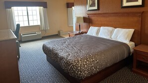 Une chambre d’hôtel avec un grand lit, un bureau et une chaise. Il y a une fenêtre avec des rideaux, une lampe fixée au mur et deux tableaux encadrés sur le mur.