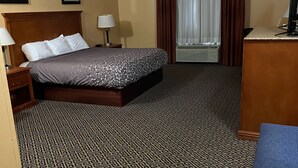 Une chambre d’hôtel comprenant un lit, un bureau, une chaise, une lampe, une fenêtre avec des rideaux et un tapis à motifs.