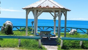 A gazebo with a metal roof and white pillars, located at a beach with a clear blue sky and ocean in the background.