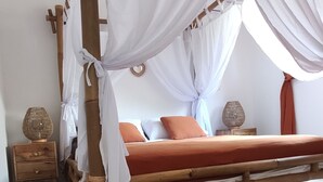 A wooden four-poster bed with white drapes, a red mattress, and a brown rug on a tiled floor.