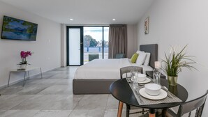 A modern hotel room with a large bed, a dining area with a table and chairs, a view through the window, and a painting on the wall.