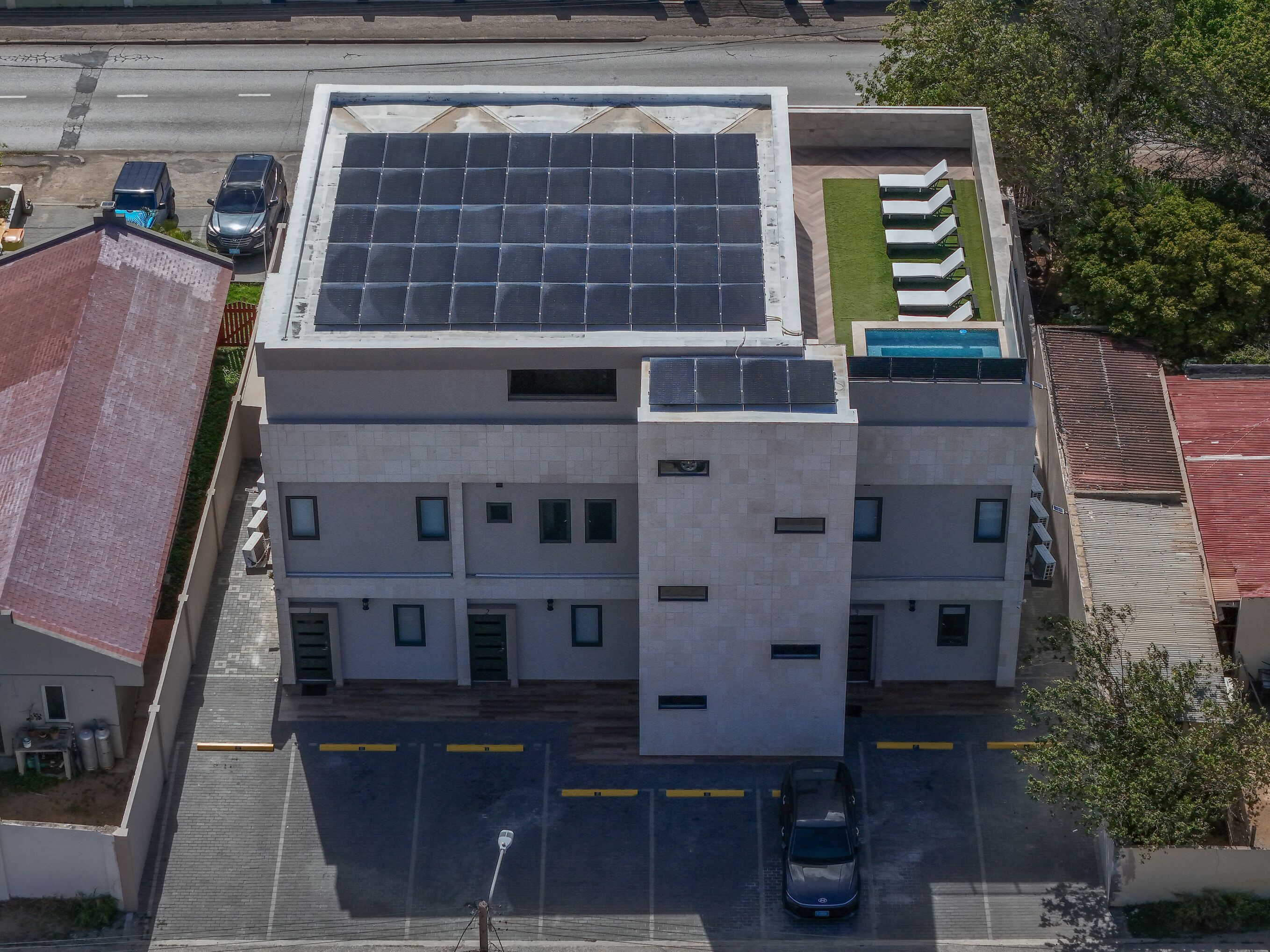 A multi-story building with a rooftop solar panel array, a parking lot, and a small green area with lounge chairs.