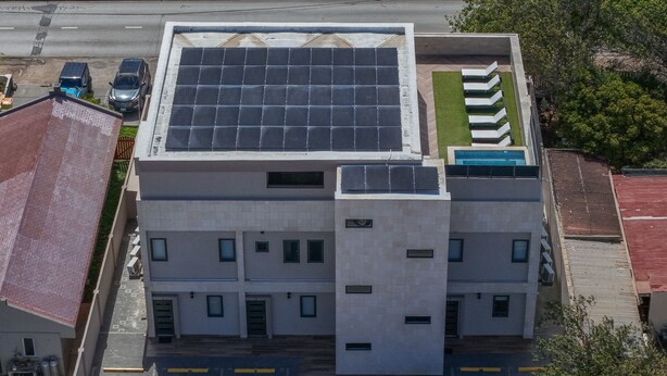 A multi-story building with a rooftop solar panel array, a parking lot, and a small green area with lounge chairs.