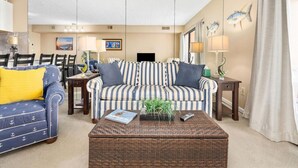 A living room with a striped sofa, a blue sofa with yellow cushions, a wicker coffee table, and a dining area with chairs and a table. - Ocean City Bayfront Ponte Vista (Ocean City)