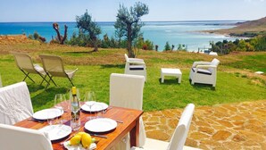 An outdoor dining area with a view of the sea, white chairs, a wooden table set for a meal, and a stone pathway. - CAMELIA by KlabHouse-2BR w/AC&Terrace-walk2beach (Sciacca)