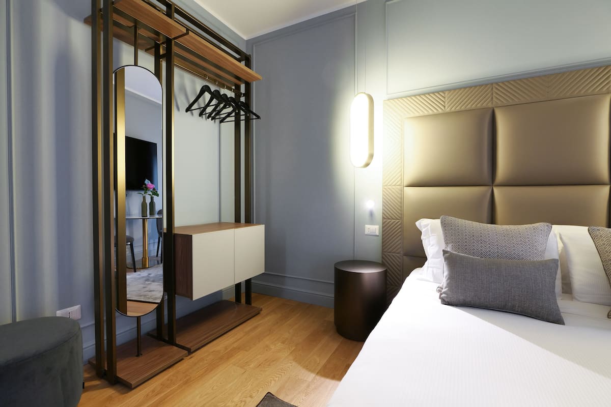 Una habitació d'hotel moderna amb sòl de fusta, un llit gran amb roba de llit gris i blanca, una tauleta de nit, un mirall amb marc de fusta i una làmpada penjada a la paret.