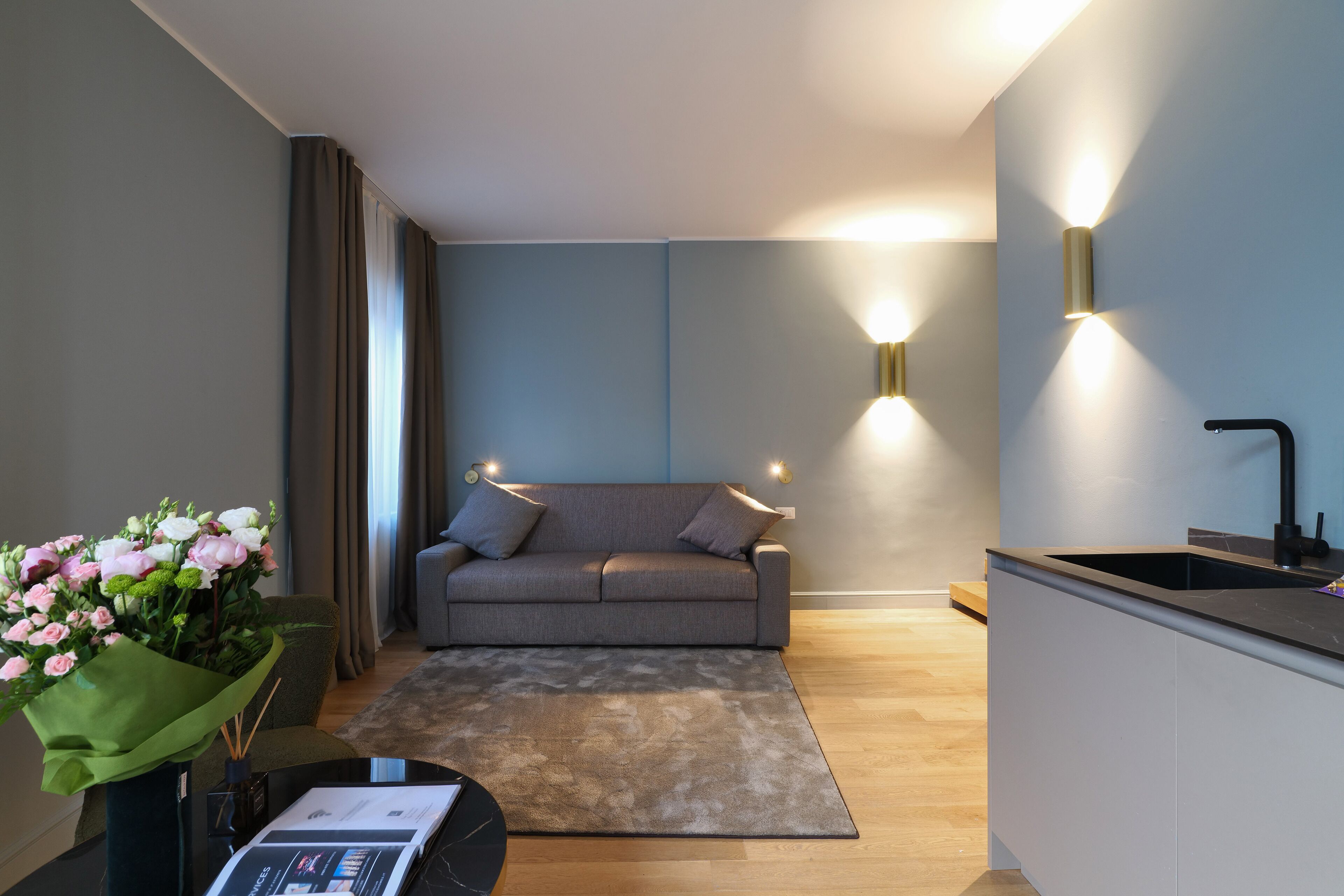 Ein modernes Wohnzimmer mit grauem Sofa, einem Couchtisch mit einer Zeitschrift, einer grünen Vase mit Blumen und einer Küchenzeile mit Spüle und Arbeitsfläche.