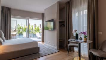 A modern bedroom with a large bed, a TV mounted on the wall, a dining table with chairs, and a balcony with a view of buildings and greenery.