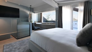 Een moderne hotelkamer met een groot bed, een glazen scheiding, een flatscreen televisie, een klein bureau en uitzicht op de stad.