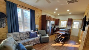 A cozy living room with a sofa and armchairs, a dining table with chairs, wooden cabinets, a refrigerator, and a kitchen area with a window. - New cabin- access to horse trails, Pilot Mtn & Hanging Rock St. Pks (Pinnacle)
