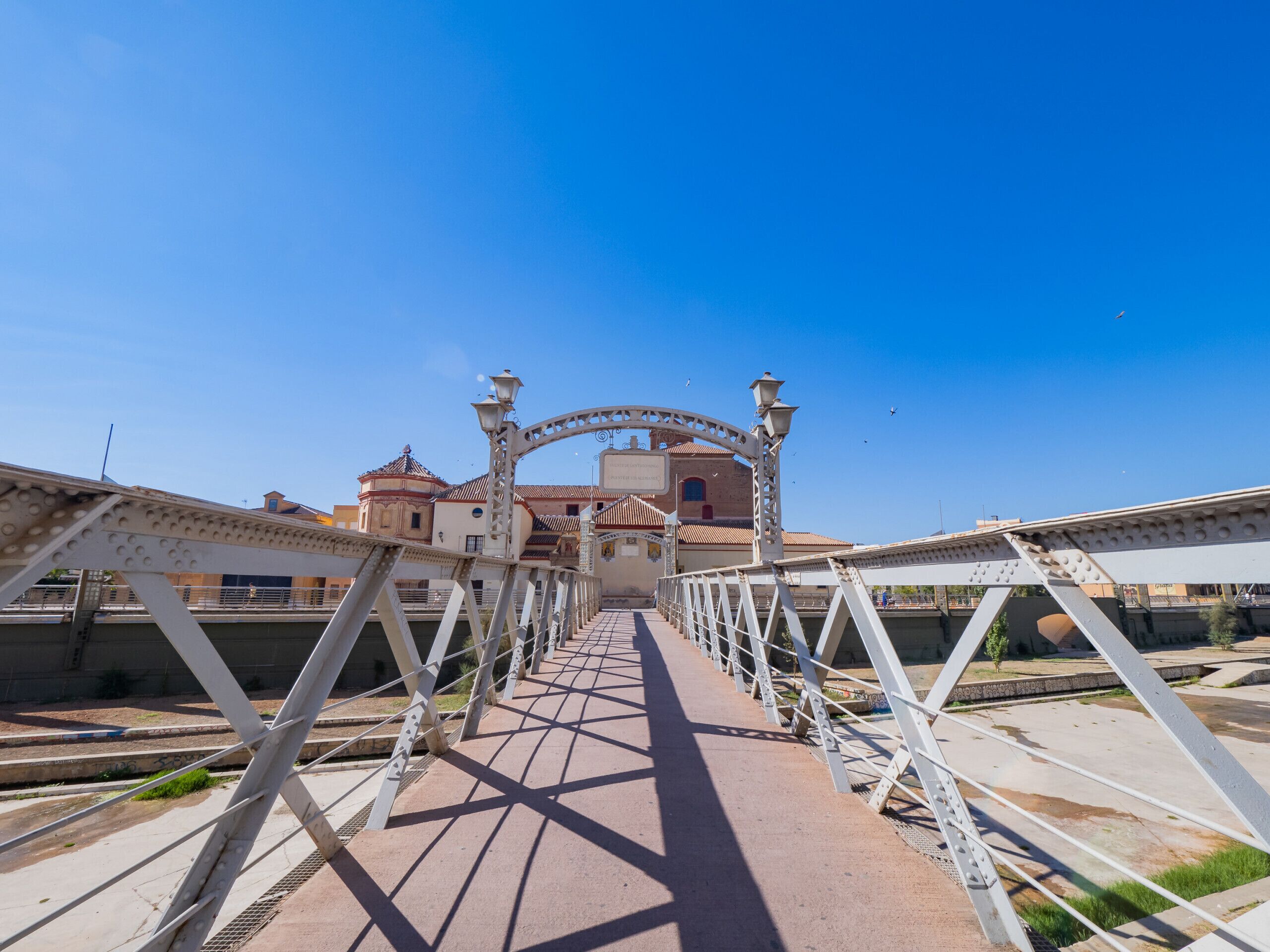 A pedestrian bridge with a metal framework and a pathway leading to a building with a dome and tiled roof.