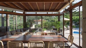 Een overdekte eethoek buiten met een houten tafel en rieten stoelen, met uitzicht op een zwembad en weelderige begroeiing.