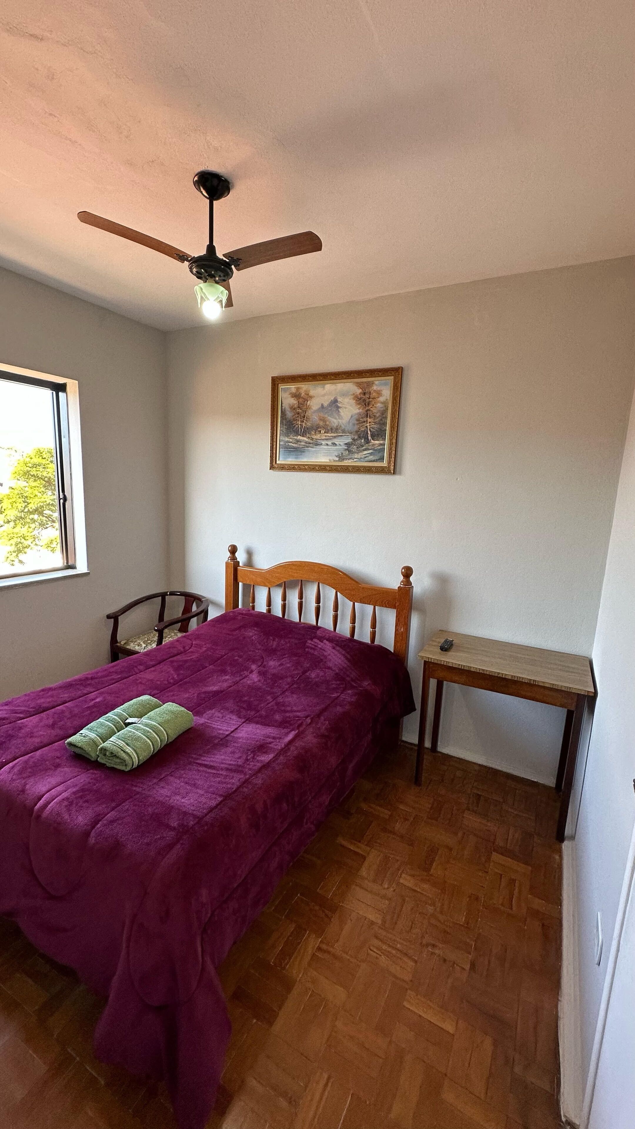 A bedroom with a wooden bed, a ceiling fan, a painting, and a small table.