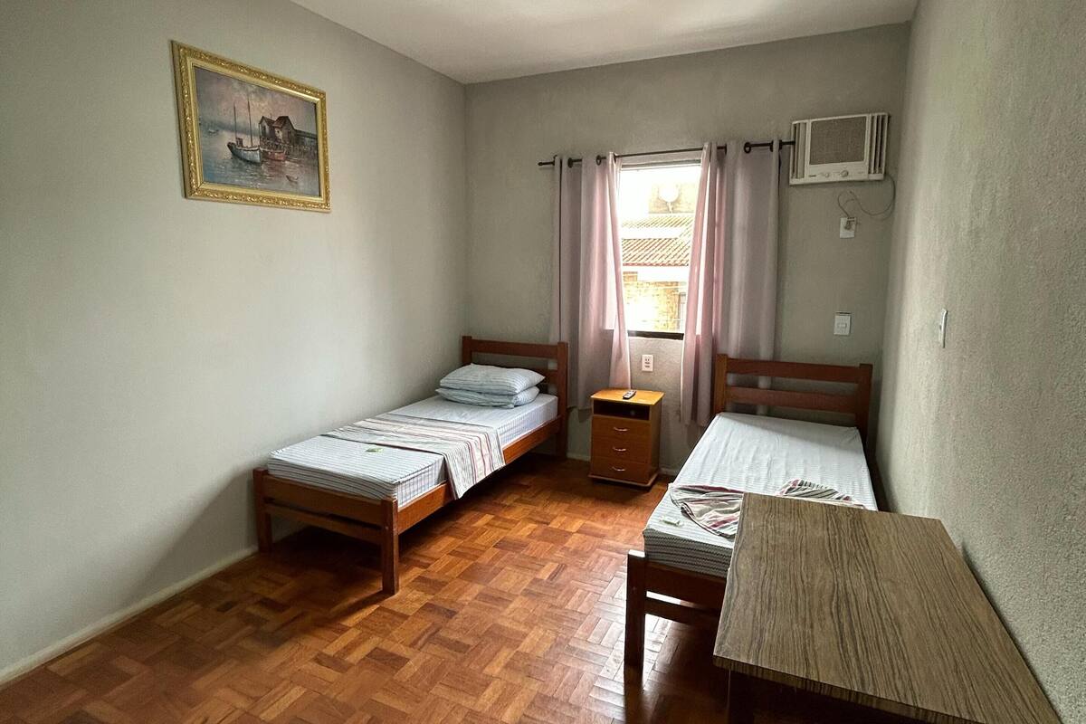 Un petit appartement d’une seule pièce, comprenant deux lits en bois, une table en bois, un tableau accroché au mur, une fenêtre avec des rideaux et un appareil de climatisation.