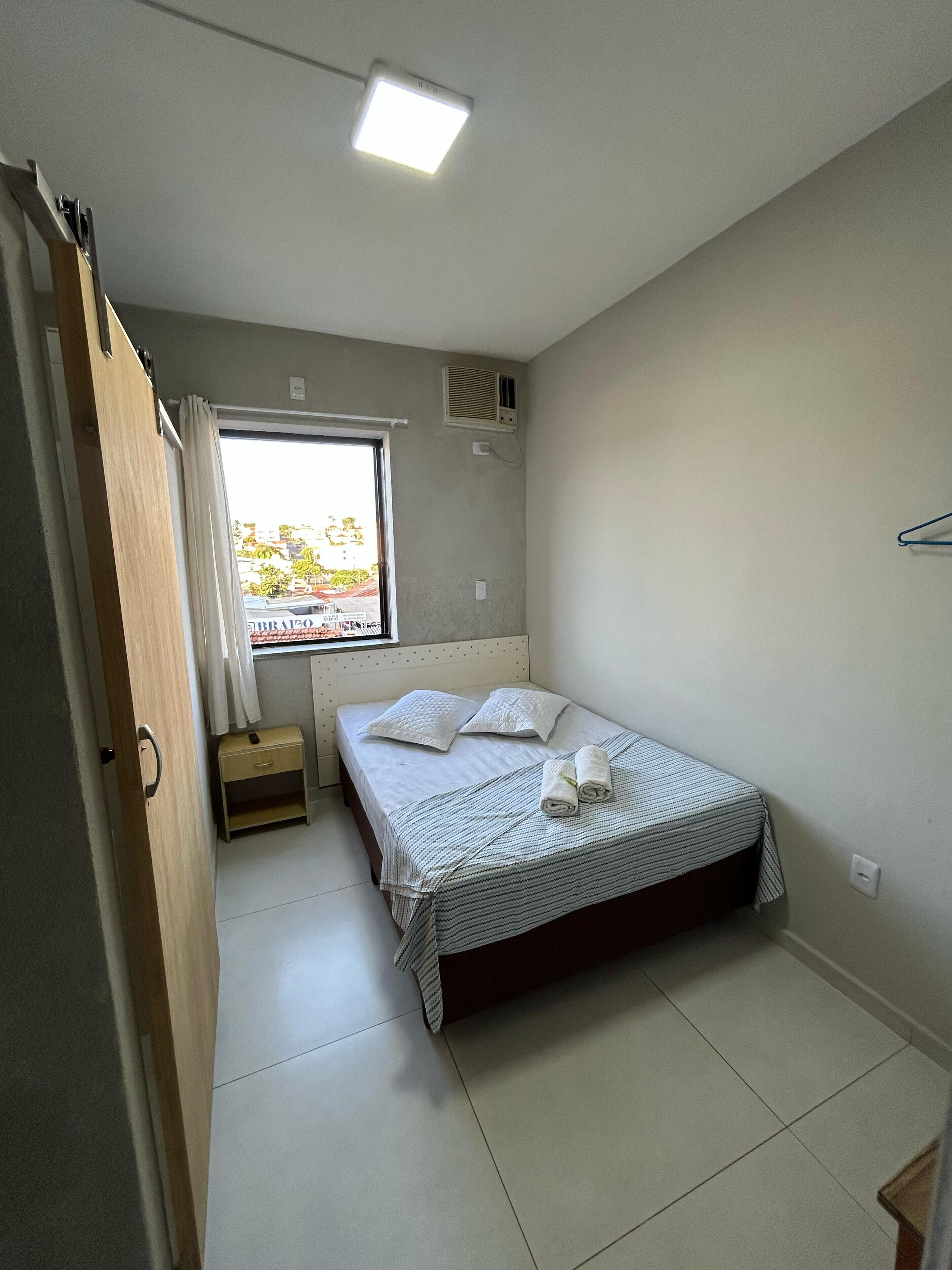 Un dormitorio pequeño y limpio con cama, mesita de noche, ventana con cortinas e iluminación en el techo.
