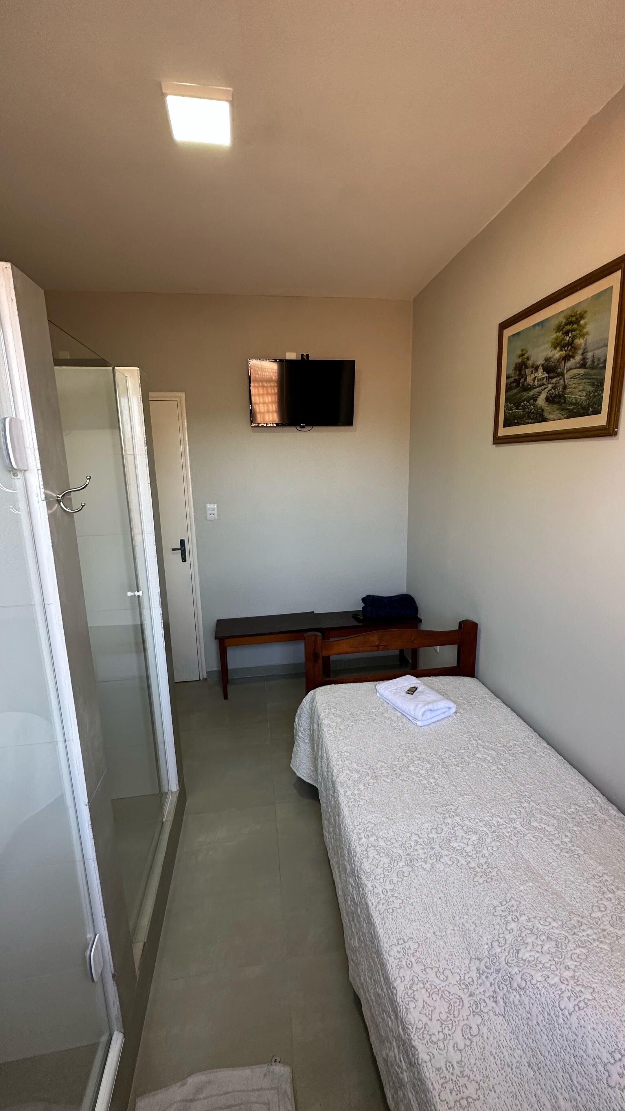 A small, well-lit room with a bed, a desk, a television mounted on the wall, and a door leading to a bathroom.