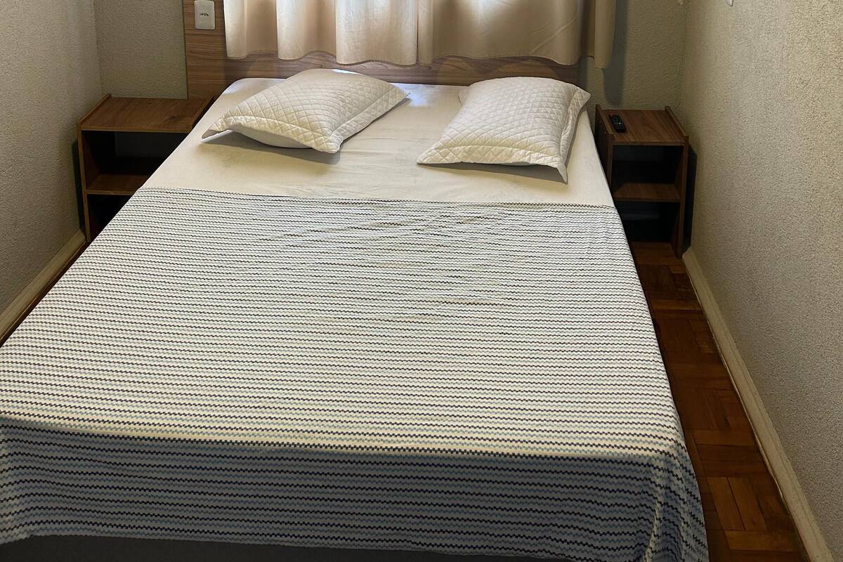 Une petite chambre à coucher, bien rangée, avec un lit, deux tables de chevet, une commode, un climatiseur fixé au mur et une fenêtre avec des rideaux.