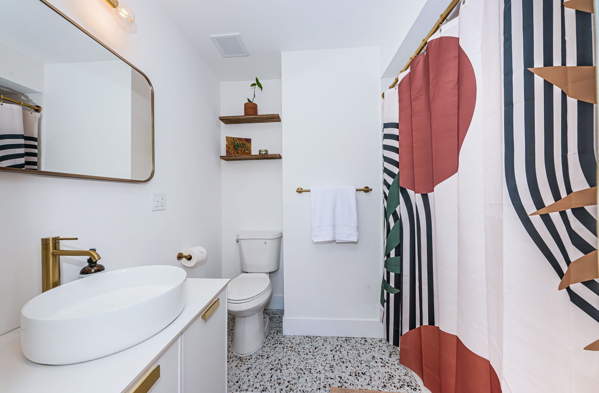 A modern bathroom with a white sink, gold faucet, and toilet. There is a shower curtain with a striped pattern and a red accent.