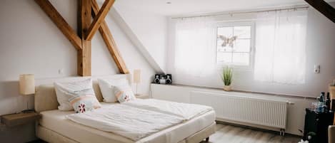Un dormitorio con cama, mesitas de noche, una ventana con cortinas, un radiador y una pequeña mesa con una planta.