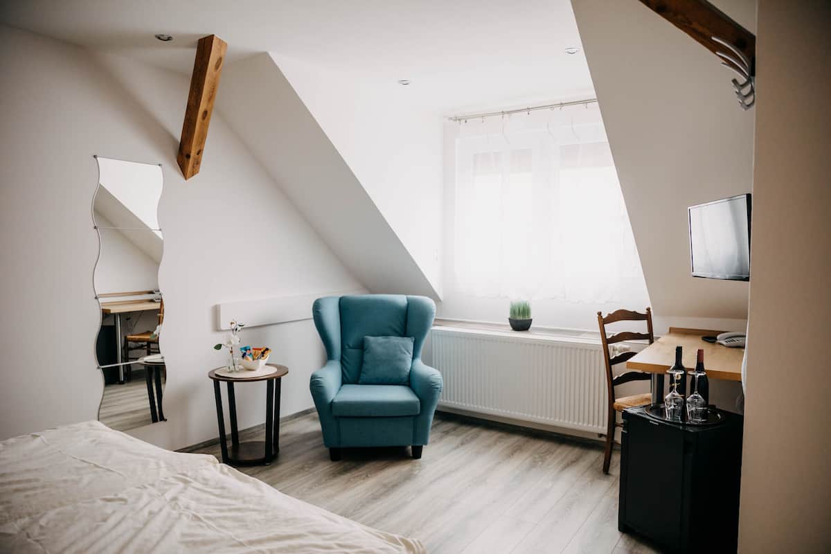 A modern bedroom with a sloped ceiling, a teal armchair, a wooden desk, a bed, and a television.