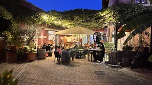 Un espace extérieur aménagé pour les repas, avec des tables et des chaises, entouré de plantes en pot et d’un mur en briques.