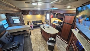 Una cocina compacta con una isla central, un comedor con mesa y asientos acolchados, y un televisor montado en la pared.