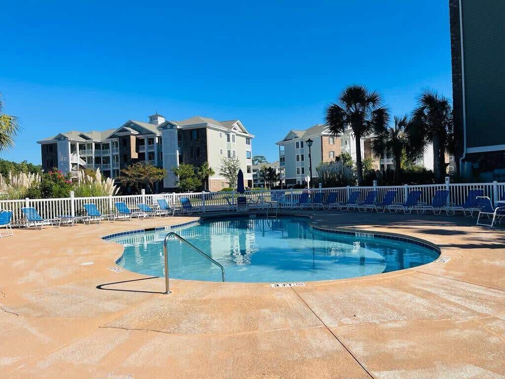 A pool area with blue lounge chairs and a pool, surrounded by a white fence, with buildings in the background.