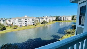 A view of a residential area with a body of water, multiple apartment buildings, and a balcony railing. - Amazing Pond View condo + pool at MyrtleWoods (Myrtle Beach)